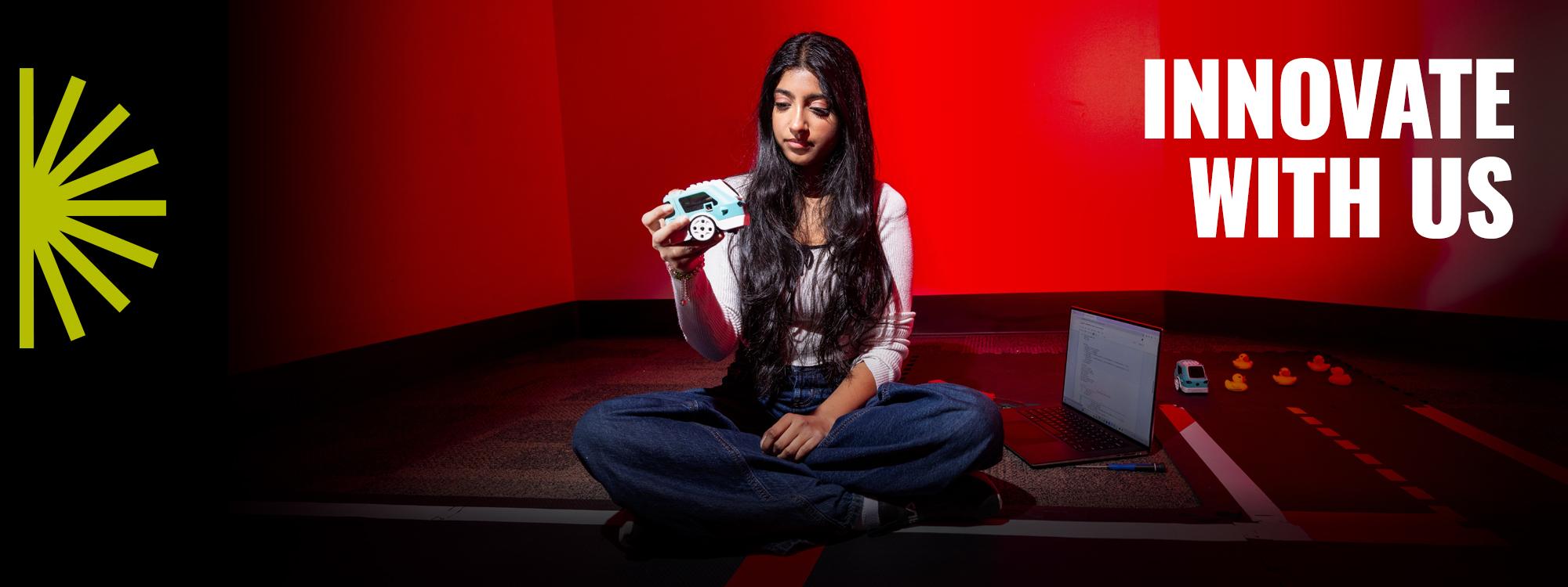 Innovate with Us text. A seated student on the floor holding a prototype for a car.